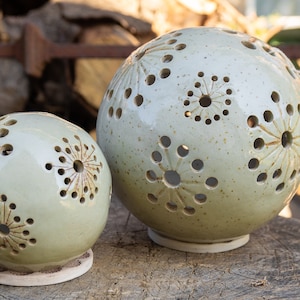 Könnte beinhalten: Zwei kugelförmige Kerzenhalter aus Keramik mit hellgrüner Glasur. Die Kugeln haben ausgeschnittene florale Designs. Die Kerzenhalter stehen auf einer Holzoberfläche, mit einem Hintergrund aus Brennholz.