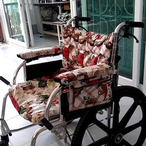Puede incluir: Silla de ruedas plateada con ruedas y reposabrazos negros. El asiento y el respaldo presentan un estampado floral con detalles en rojo y marrón. La silla está frente a una puerta de cristal.