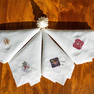 May include: Four white linen napkins with embroidered designs. The designs include a cup of coffee, a gift box with a bow, a Christmas tree, and a red sweater.