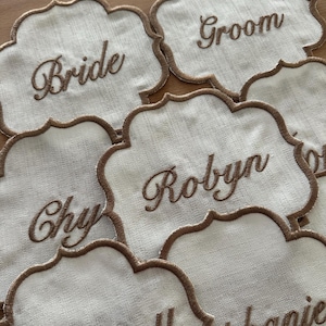 Può includere: Segnaposto ricamati bianchi con bordo smerlato e cuciture marroni. Le carte presentano nomi e titoli come "Bride" e "Groom" in elegante scrittura, perfetti per decorazioni di matrimoni o eventi.