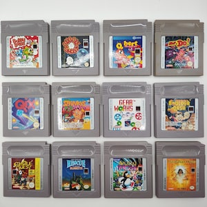 Puede incluir: Una colección de doce cartuchos de juegos grises de Nintendo Game Boy. Las etiquetas de los juegos presentan gráficos coloridos y títulos como "Bubble Bobble", "Q*bert" y "Boulder Dash". Los cartuchos están dispuestos en una cuadrícula.