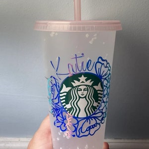 Op de afbeelding: Een doorzichtige herbruikbare beker van plastic met een roze deksel en een rietje. De beker heeft een groen Starbucks-logo met een bloemmotief eromheen. De naam "Katie" staat in blauw op de beker.