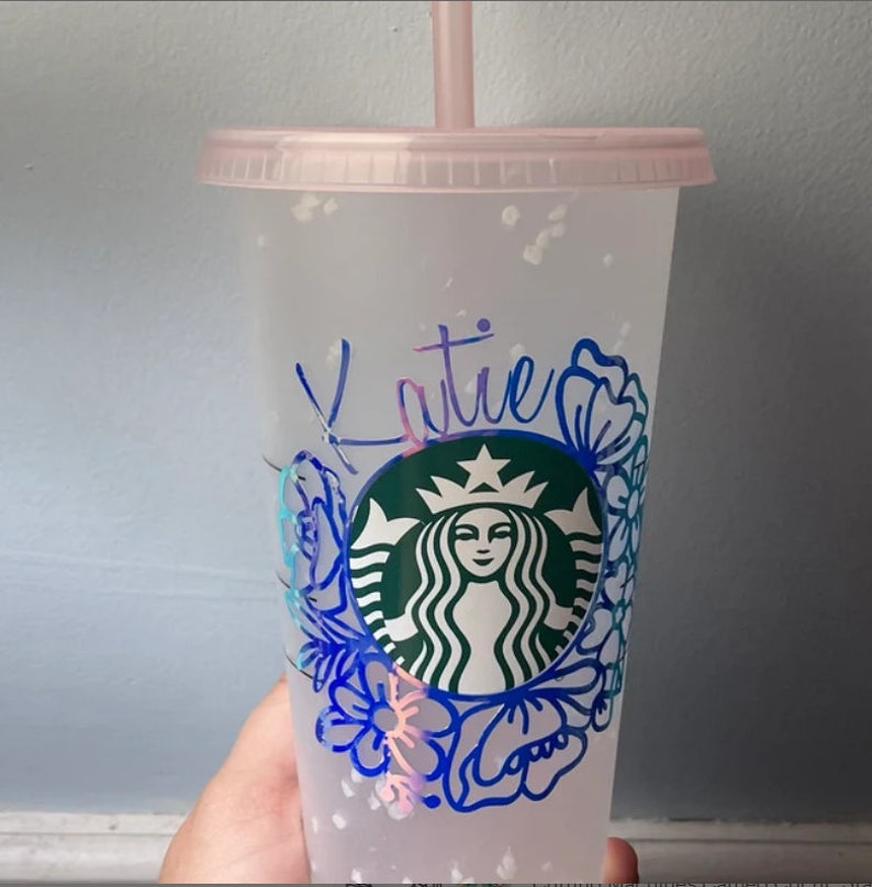 Op de afbeelding: Een doorzichtige herbruikbare beker van plastic met een roze deksel en een rietje. De beker heeft een groen Starbucks-logo met een bloemmotief eromheen. De naam "Katie" staat in blauw op de beker.
