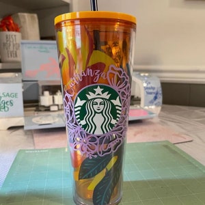 Op de afbeelding: Een doorzichtige herbruikbare beker met een geel deksel en een rietje. De beker heeft een bloemmotief met een Starbucks-logo en het woord "Carranza" in cursief.