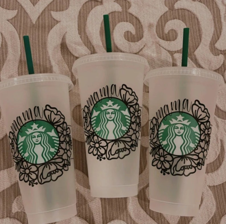 Op de afbeelding: Drie doorzichtige plastic bekers met zwarte en groene ontwerpen. Elke beker heeft een groene rietje en een zwart ontwerp dat "mama" zegt met een bloemenpatroon en een groen Starbucks-logo.