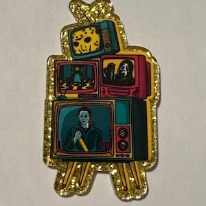 May include: A glittery gold pin shaped like a stack of vintage televisions. Each screen displays a horror movie scene, including Jason Voorhees, Ghostface, and Michael Myers. The pin has a gold glitter border and is a unique accessory.
