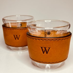 May include: Two clear glass cups with a brown leather sleeve. The leather sleeves have a stitched design and the letter "W" embossed on the front. The cups are designed with a double-walled construction.