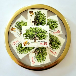 Pu&ograve; includere: Una collezione di francobolli vintage "White Oak USA 15&cent;" disposti in una cornice circolare dorata. I francobolli presentano un'illustrazione di un albero verde con il testo "WHITE OAK" e "USA 15&cent;".