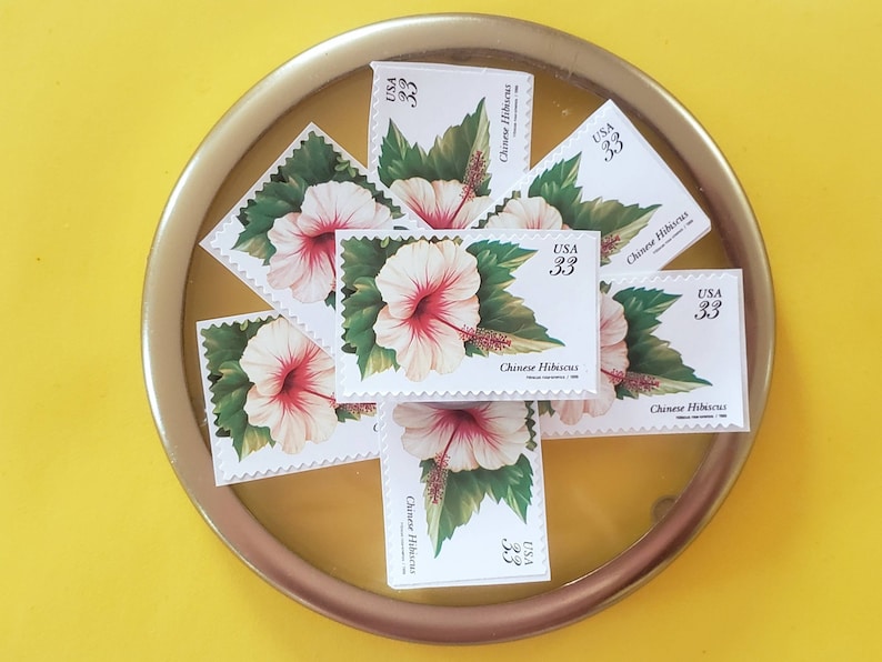 May include: A collection of eight vintage Chinese Hibiscus postage stamps, each featuring a white flower with a red centre and green leaves. The stamps are arranged in a circular pattern on a gold-coloured surface. Each stamp has the text "USA 33" and "Chinese Hibiscus" printed on it.