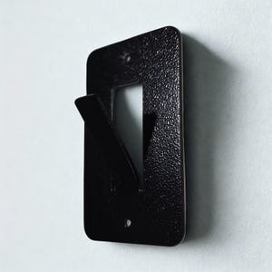 May include: A black metal wall hook with a textured finish. The hook has a rectangular backplate with a cutout and a projecting hook. The hook is mounted on a white wall.