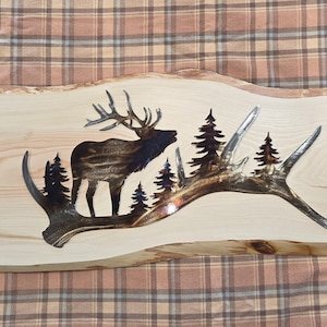 May include: Wooden wall art featuring a silhouette of an elk with antlers, set against a backdrop of pine trees. The elk and trees are dark brown, and the antlers are silver. The art is mounted on a light-colored wooden plank.
