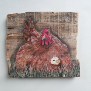 Può includere: Opera d'arte dipinta a mano su tavola di legno raffigurante una gallina e il suo pulcino. La gallina è marrone con cresta rossa, e il pulcino è giallo. L'opera è ambientata su uno sfondo di erba verde e venature del legno.