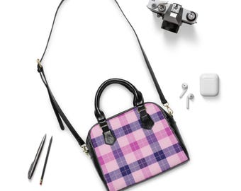 May include: A pink, purple, and navy plaid pattern satchel bag with a black handle and strap. The bag is set on a white background with a camera, earbuds, and a pencil.