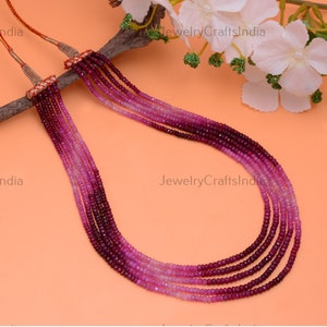 May include: A multi-strand necklace made of pink and red gemstone beads. The beads are graduated in size, with the larger beads at the bottom of the necklace. The necklace is strung on a gold cord.