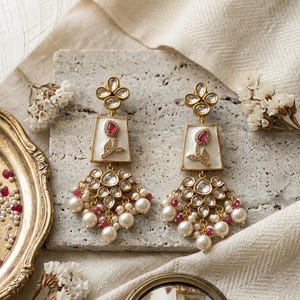 Könnte beinhalten: Goldfarbene Ohrringe mit floralem Design. Jeder Ohrring hat eine rechteckige, weiße Emaillebasis mit einem roten Edelstein und einem Blumenmotiv. Die Ohrringe haben eine Ansammlung von Perlen und roten Edelsteinen.