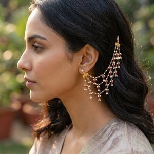 Puede incluir: Perfil de una persona con un pendiente de oreja dorado con hebras colgantes de cuentas blancas y rojas. El pendiente de oreja está unido a un pequeño pendiente de botón. La persona tiene el pelo oscuro y lleva una blusa clara.