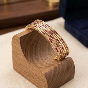 May include: Gold bangles with alternating rows of red gemstones and clear crystals, displayed on a wooden stand. The jewelry is set against a soft, neutral background, highlighting the intricate design and sparkle of the bracelets.