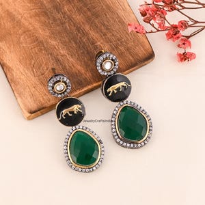 Può includere: Orecchini pendenti con un ciondolo circolare nero con un disegno di tigre dorata, una pietra verde a forma di goccia e un bordo color argento. Gli orecchini sono appoggiati su una superficie di legno con fiori rosa.