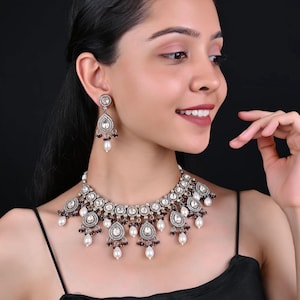 Puede incluir: Un conjunto de collar y pendientes plateados. La joyería presenta colgantes en forma de lágrima con piedras transparentes, pequeños acentos rojos y perlas blancas colgantes. El collar lo lleva una persona que lleva una camiseta negra.