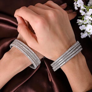 Puede incluir: Un conjunto de cinco pulseras de plata con filas de piedras de circonita cúbica transparentes. Las pulseras están apiladas unas sobre otras y se llevan en la muñeca.