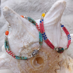 May include: Two beaded bracelets with silver accents. One bracelet has turquoise, red, and orange beads. The other bracelet has turquoise, blue, and yellow beads. Both bracelets have a silver charm in the shape of a Buddha's face.