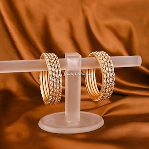 Könnte beinhalten: Ein Set aus sechs goldenen Armreifen mit klaren, facettierten Steinen. Die Armreifen werden auf einem weißen Ständer präsentiert. Der Text "jewelryCraftsIndia" ist auf dem Ständer sichtbar.
