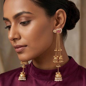 May include: Gold-toned earrings with a floral design and dangling bell-shaped charms. The earrings feature small pearls and red accents. The model is wearing a maroon top.