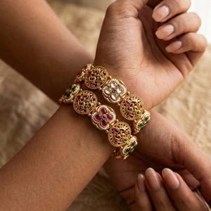 Peut inclure: Deux bracelets rigides dorés composés d'une série de maillons circulaires en filigrane. Chaque maillon est orné de petites perles blanches, de pierres rouges éclatantes et d'accents verts disposés en motifs floraux sur le métal.
