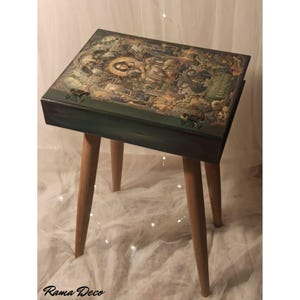 May include: A green book-shaped side table with a whimsical, vintage-inspired design. The table top features a colorful illustration of a fairy tale scene with a woman and animals. The table has four wooden legs.