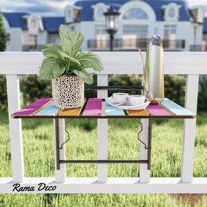 Op de afbeelding: Een kleurrijke opklaptafel met een zwart metalen frame bevestigd aan een wit hek. De tafel heeft een roze, blauwe en gele gestreepte bovenkant en is versierd met een plant in een pot en twee kopjes koffie op een bord.