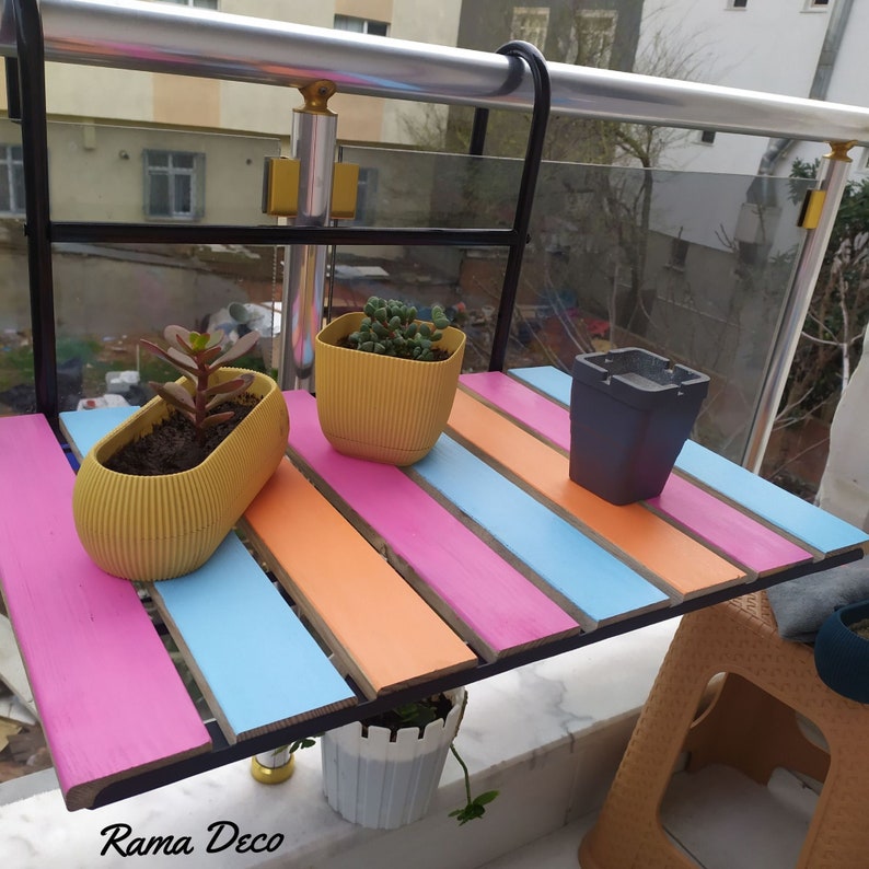 Op de afbeelding: Een zwarte metalen plantenstandaard met een kleurrijk houten blad. Het blad is geschilderd in strepen van roze, blauw en oranje. Er staan drie potplanten op de standaard, twee in gele potten en &eacute;&eacute;n in een grijze pot. De standaard is bevestigd aan een railing.