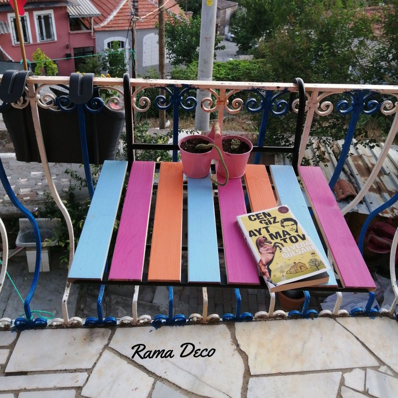 Op de afbeelding: Een kleurrijk houten bankje met blauwe, roze en oranje strepen staat op een balkon met een smeedijzeren railing. Een boek getiteld "Cen Giz AYT MA TOv" door "Elveda Gulsari" ligt op het bankje. Twee roze bloempotten met groene planten hangen aan de railing.
