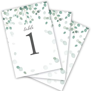 May include: Set of three white table number cards with a green eucalyptus leaf design. The cards have the text "table 1" printed on them.