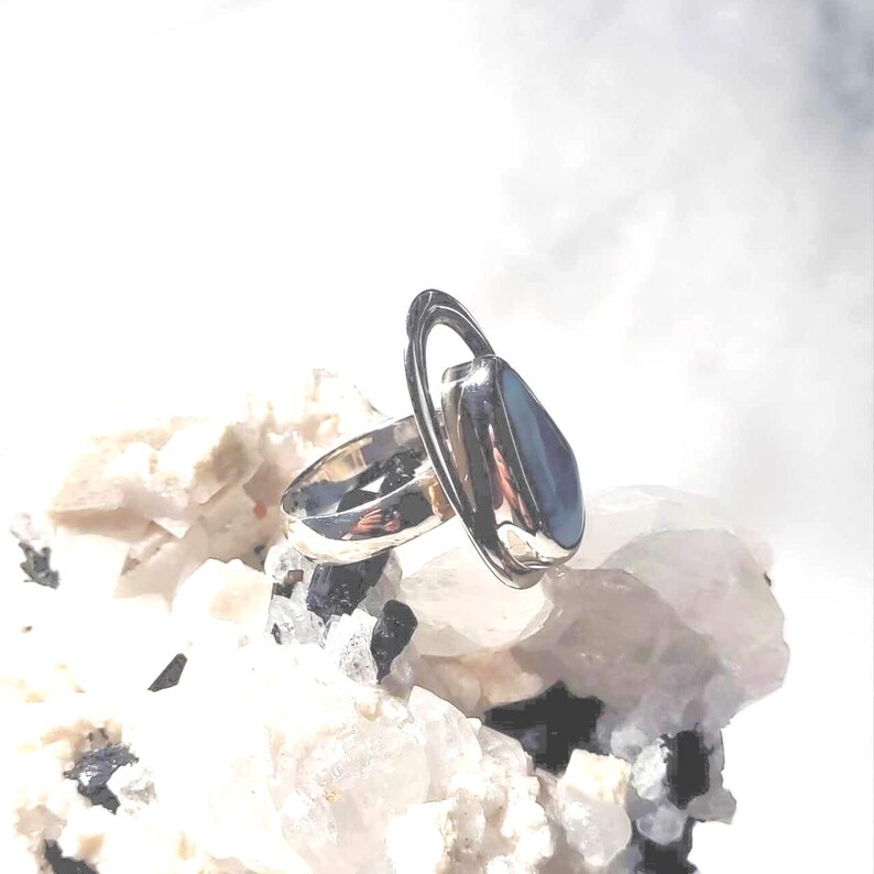 May include: A silver ring with a large, oval, blue-grey stone. The ring has a unique design with a loop encircling the stone. The ring is displayed on a bed of white and black rocks, creating a striking contrast.