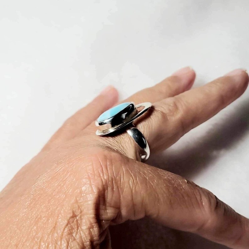 May include: A silver ring featuring a light blue oval-shaped stone. The ring has a unique design with a circular accent around the stone. The ring is shown on a hand.