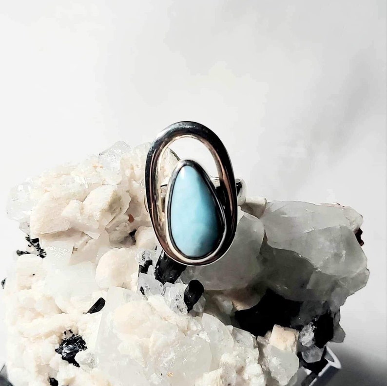 May include: A silver ring featuring a light blue, teardrop-shaped stone set within an open oval frame. The ring is displayed on a white and gray mineral formation, creating a contrast of textures and colors. A piece of jewelry.