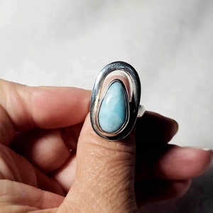 May include: A silver ring featuring a teardrop-shaped, light blue gemstone. The stone is set within a silver oval frame. The ring is shown on a finger against a white background.