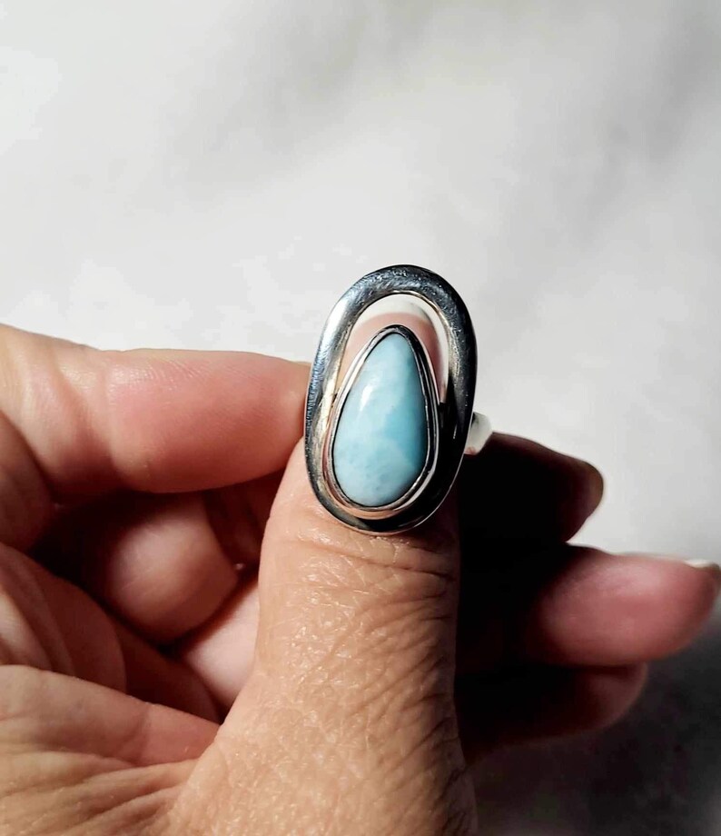 May include: A silver ring featuring a teardrop-shaped, light blue gemstone. The stone is set within a silver oval frame. The ring is shown on a finger against a white background.