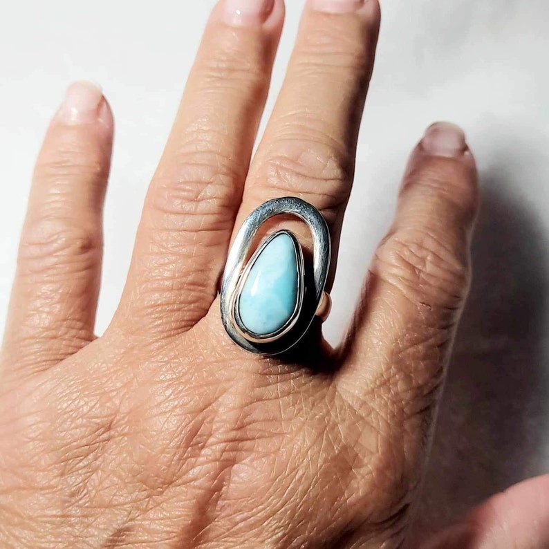 May include: A silver ring featuring a teardrop-shaped, light blue gemstone. The gemstone is set within a silver frame, and the ring has a unique, open design. The ring is shown on a person's finger.