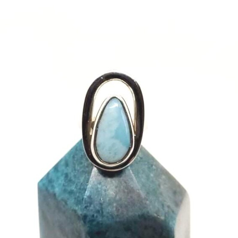 May include: A silver ring with a teardrop-shaped, light blue gemstone. The gemstone is set within a silver oval frame. The ring is displayed on a blue-green, geometric object against a white background.