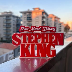 Może przedstawiać: Czerwony znak 3D z napisem "Stephen King" białymi literami. Nad nazwiskiem znajduje się mniejszy biały znak z tekstem "Your Name's Library".