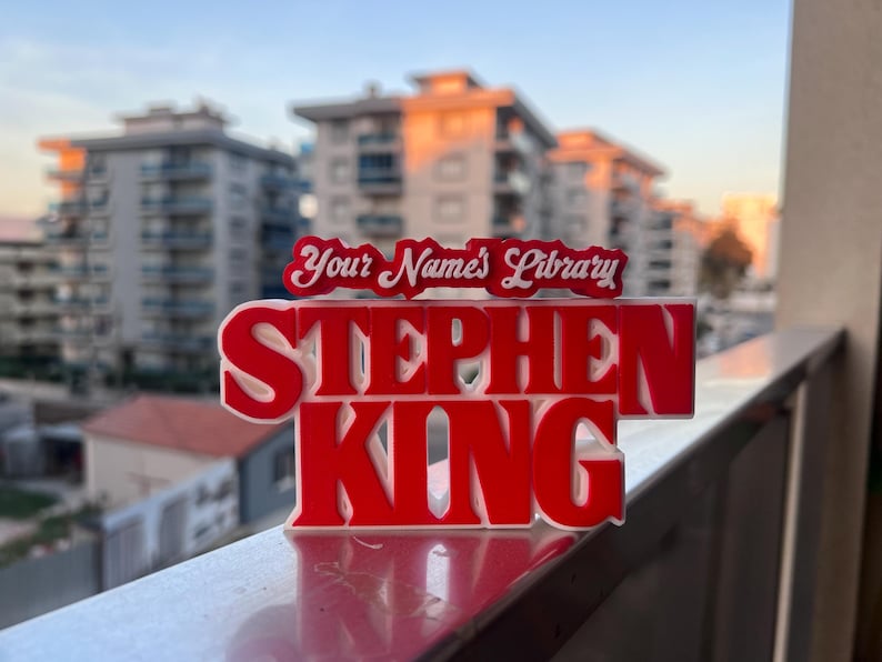 Może przedstawiać: Czerwony znak 3D z napisem "Stephen King" białymi literami. Nad nazwiskiem znajduje się mniejszy biały znak z tekstem "Your Name's Library".