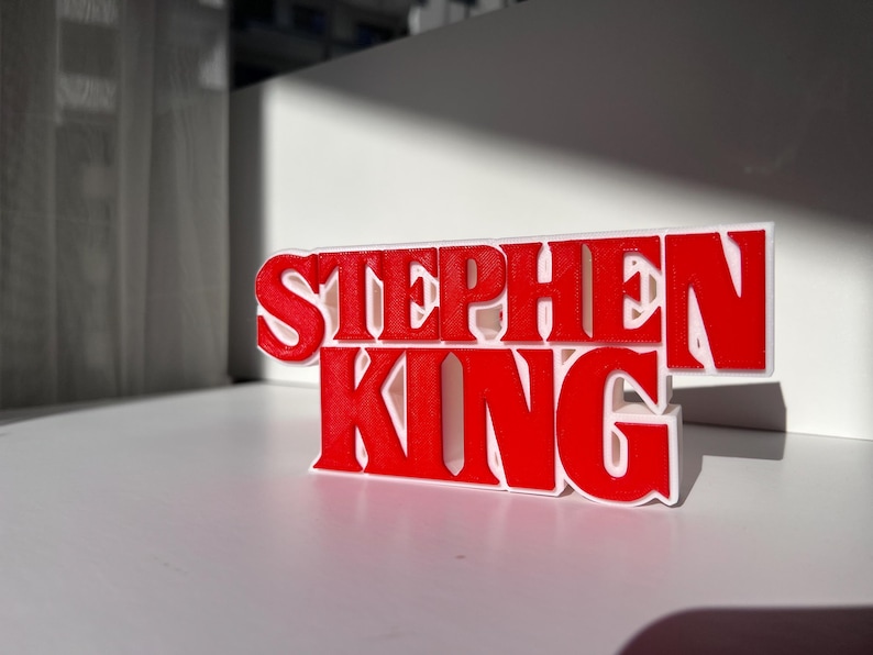 Może przedstawiać: Tr&oacute;jwymiarowy znak z napisem "Stephen King" w czerwonych literach na białym tle.