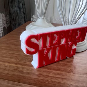 Może przedstawiać: Biały znak 3D z napisem "Stephen King" czerwonymi literami.