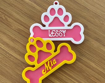 May include: Two pink and yellow dog bone-shaped tags with paw print designs. The top tag is white with the name "Lessy" in yellow lettering. The bottom tag is yellow with the name "Mia" in white lettering.