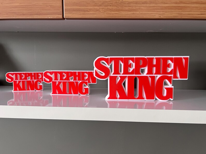 Może przedstawiać: Trzy czerwone, tr&oacute;jwymiarowe tabliczki z napisem "Stephen King" w r&oacute;żnych rozmiarach. Największa tabliczka znajduje się z tyłu, a dwie mniejsze z przodu.