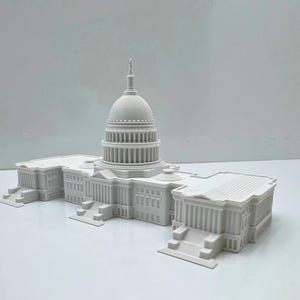May include: A 3D-printed model of the United States Capitol Building. The model is white and features detailed architectural elements, including the dome, columns, and steps. The building is a miniature replica of the iconic landmark.
