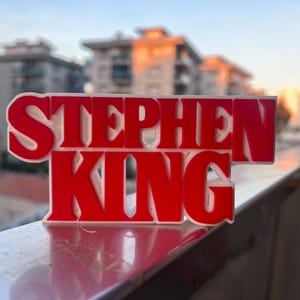 Może przedstawiać: Tr&oacute;jwymiarowa tabliczka z napisem "Stephen King" w czerwonych literach z białymi obramowaniami. Tabliczka leży na srebrnej balustradzie.