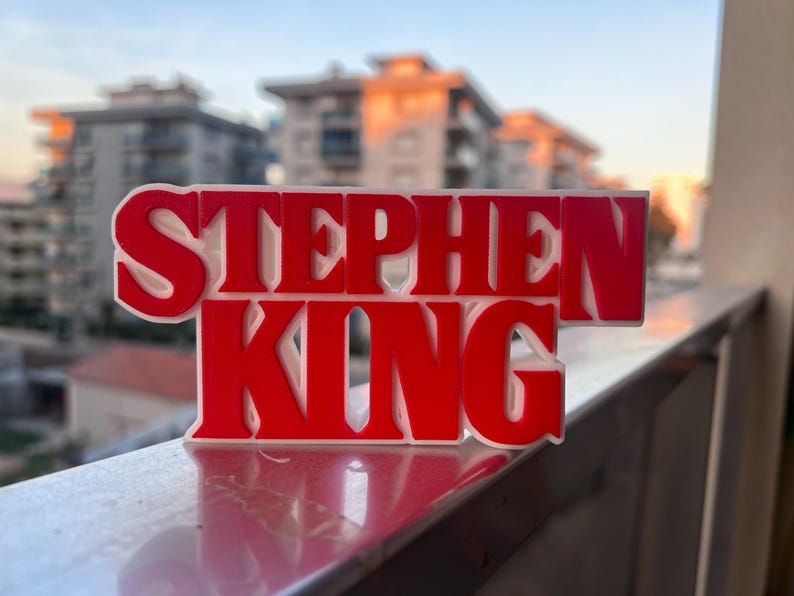 Może przedstawiać: Tr&oacute;jwymiarowa tabliczka z napisem "Stephen King" w czerwonych literach z białymi obramowaniami. Tabliczka leży na srebrnej balustradzie.
