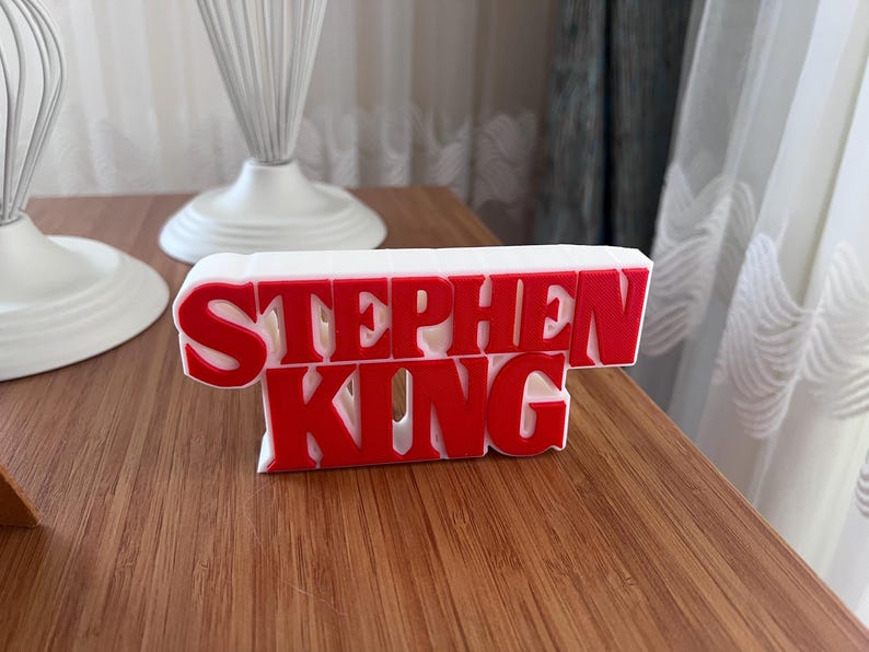Może przedstawiać: Biały znak 3D z napisem "Stephen King" czerwonymi literami.